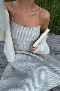 Femme en robe rayée et cardigan blanc tenant un livre, assise sur des planches de bois avec de l'herbe verte en arrière-plan.