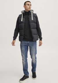 Chaleco acolchado negro con capucha, con cordones blancos, llevado sobre un suéter oscuro y jeans azules, combinado con zapatillas negras.