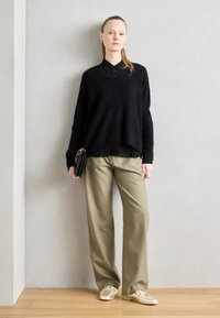 Un pull noir en peluche superposé à une chemise à col noire, accompagné d'un pantalon large vert olive et de chaussures décontractées métalliques. Tient une pochette noire.