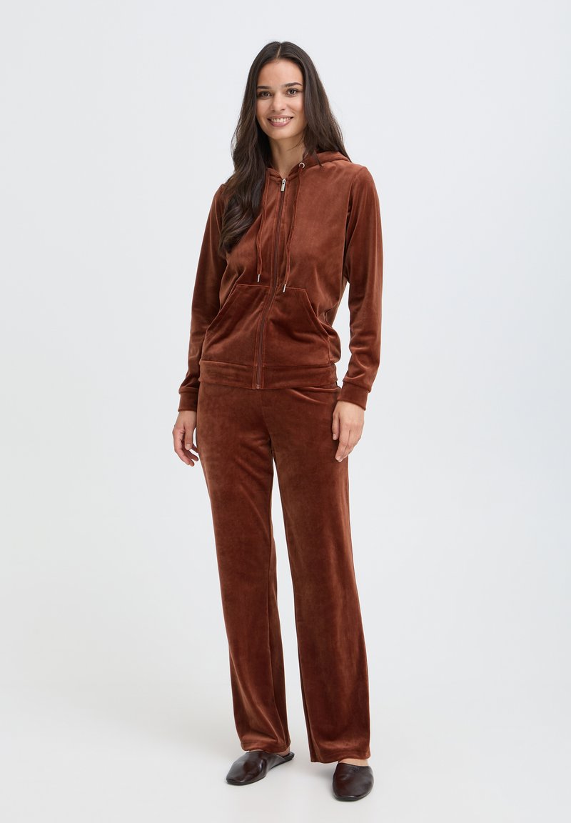 Femme debout et souriante, portant un sweat à capuche zippé en velours marron et un pantalon large assorti avec des chaussures sombres sans lacets.