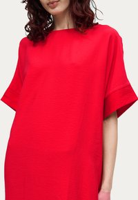 Femme portant une robe rouge vif à manches courtes amples avec un décolleté bateau, posant devant un arrière-plan uni.