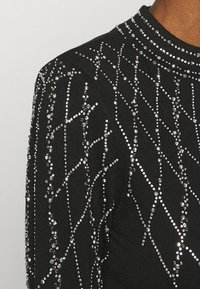 Pull noir présentant un motif texturé avec des strass argentés disposés en motif croisé le long des manches et du col.