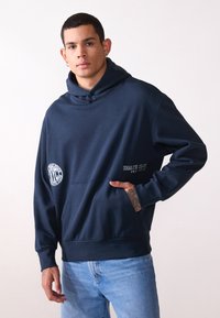 Navy blue hooded sweatshirt made of soft fabric, features a front pocket, embroidered logos, and ribbed cuffs. Casual fit with subtle text details.