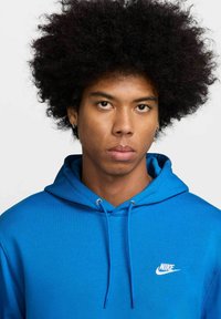 Sudadera azul con cordón, presenta un pequeño logo blanco de Nike en el pecho. La tela parece suave y texturizada, con un corte holgado.
