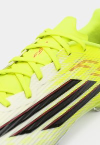 Φωτεινό κίτρινο ποδοσφαιρικό παπούτσι με υφή στην επάνω πλευρά, λευκές και μαύρες λεπτομέρειες, λεπτά κορδόνια και αεροδυναμικό σχεδιασμό για ευελιξία.