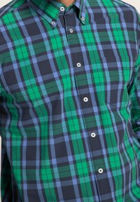 Mężczyzna ubrany w zieloną, niebieską i czarną koszulę w kratę z zapiętym kołnierzykiem i kieszenią na piersi, widoczny tors bez głowy.