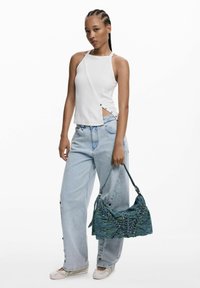 Fonott hajú nő, fehér ujjatlan fels�őt és világoskék, bő nadrágot viselve, kék mintás táskával a kezében és fehér cipőben.