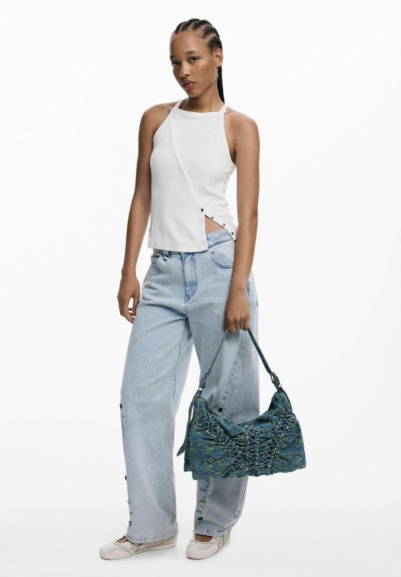 Fonott hajú nő, fehér ujjatlan felsőt és világoskék, bő nadrágot viselve, kék mintás táskával a kezében és fehér cipőben.