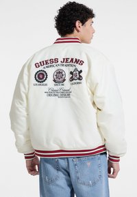 Weiße Bomberjacke mit rot-blauen gestreiften Rippbündchen und Kragen. Mit aufgestickten Logos und Text auf der Rückseite.