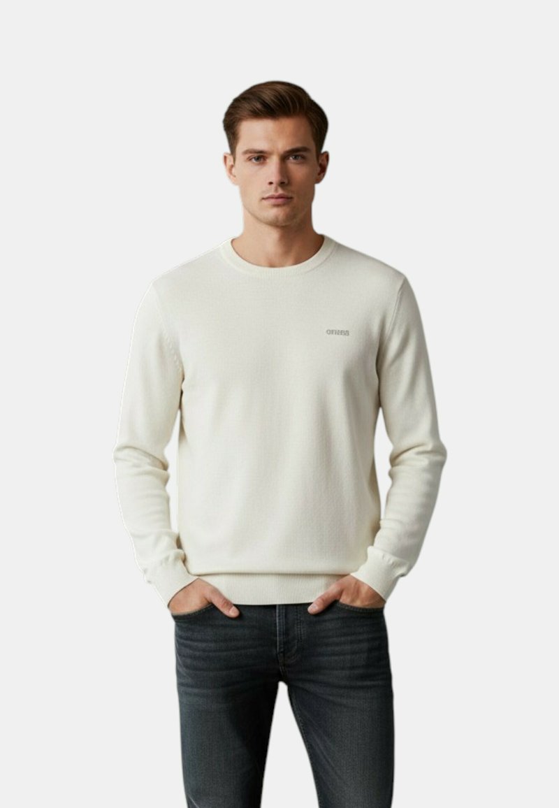 Jeune homme portant un pull crème à manches longues avec les mains dans les poches, associé à un jean gris foncé, debout devant un fond uni.
