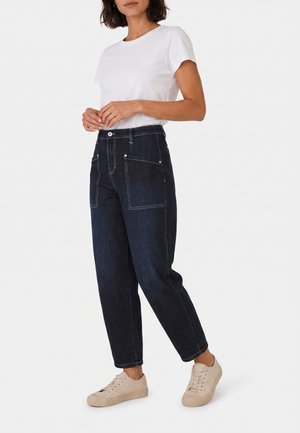 Femme portant un t-shirt blanc à manches courtes, un jean foncé taille haute avec de grandes poches avant, et des baskets beige clair.