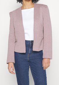 Kvinna i en mauvefärgad strukturerad blazer över en vit topp och högmidjade blå jeans, som står mot en enfärgad ljus bakgrund.