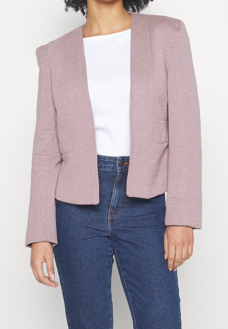 Kvinna i en mauvefärgad strukturerad blazer över en vit topp och högmidjade blå jeans, som står mot en enfärgad ljus bakgrund.