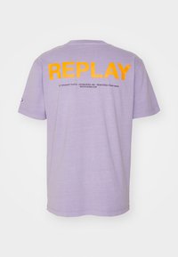 Levandulové tričko s krátkým rukávem, na zádech velký oranžový nápis "REPLAY" a menší černý text pod ním, vystavené na bílém pozadí.