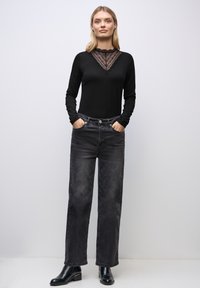 Schwarzes langärmliges Oberteil mit Spitzenakzent am Ausschnitt, kombiniert mit hoch taillierten, dunkelgrauen weiten Jeans und schwarzen Stiefeletten.