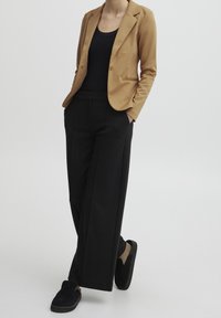 Blazer marrón claro con solapas de pico, camiseta negra sin mangas y pantalones anchos negros. Calzado de ante negro tipo slip-on. Ajuste suave y entallado.