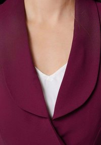 Femme portant un blazer prune profond sur un top blanc en V, révélant le décolleté et la zone supérieure de la poitrine.