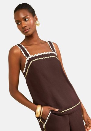 Femme portant un haut marron sans manches et un pantalon avec une bordure blanche ondulée, de grandes boucles d’oreilles dorées et deux bracelets dorés, posant devant un fond clair.