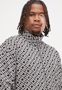Homme avec des cheveux tressés de longueur moyenne portant une veste zippée noire et blanche à motifs devant un fond uni.