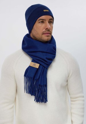 Beanie blu in lana e sciarpa abbinata con frange. La sciarpa presenta un'etichetta in pelle. Il modello indossa un maglione a coste crema. Design semplice e versatile.