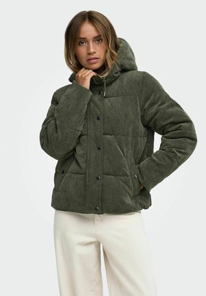 Femme portant une veste matelassée en velours côtelé vert olive avec une capuche, debout, une main dans la poche et l'autre touchant le col de la veste.