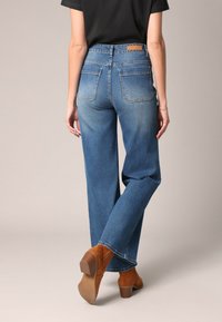 Jean bleu en denim avec une taille haute, des jambes droites et deux poches arrière. Porté avec des bottines à talons bruns.