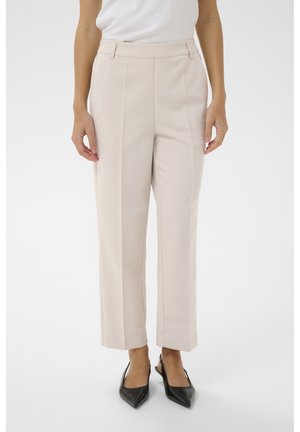 Nő, aki bézs, magas derekú, elején hajtással ellátott szabott nadrágot visel, valamint fekete, hegyes orrú, pántos magassarkú cipőt fehér háttér előtt.