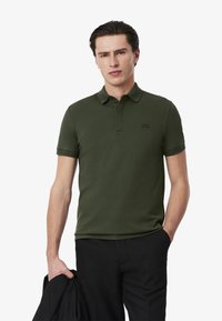 Polo shirt verde scuro con maniche corte, colletto e un piccolo logo. Il tessuto appare liscio e attillato, indossato con pantaloni scuri e giacca.