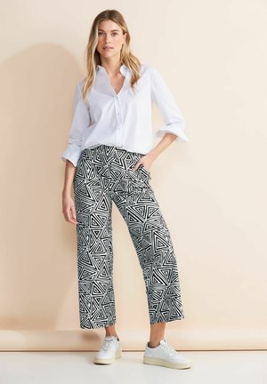 Witte button-up shirt met opgerolde mouwen, gecombineerd met zwart-wit gestreepte wijdgelegde broek met geometrische driehoekpatronen. Witte sneakers.