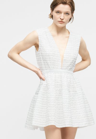 Γυναίκα με κοντά μαλλιά που φορεί ένα λευκό φόρεμα χωρίς μανίκια με υφή, βαθύ V λαιμόκοψη και φουσκωτή φούστα, τα χέρια της ακουμπούν στους γοφούς της.