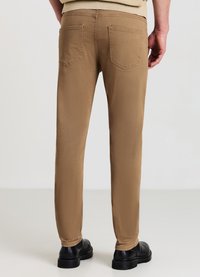 Tannfarbene Slim-Fit-Hosen mit fünf Taschen, sichtbaren Nähten und glatter Textur. Getragen mit schwarzem Schuhwerk. Rückansicht zeigt die Details des Bundes.