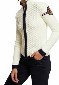 White knitted sweater with a cable pattern, navy blue accents at the collar and cuffs, and a badge on the upper arm. Zipper closure.