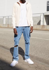 Persona che indossa una felpa con cappuccio beige chiaro con cerniera, t-shirt bianca, jeans blu strappati a un ginocchio e sneakers bianche, in piedi sul marciapiede.