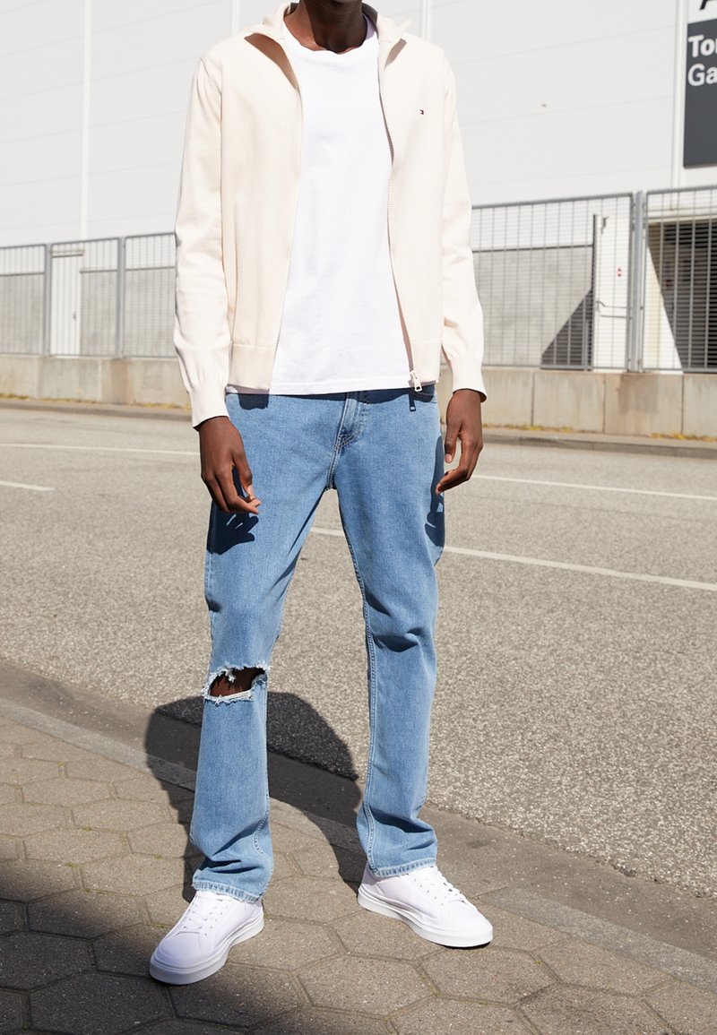 Persona che indossa una felpa con cappuccio beige chiaro con cerniera, t-shirt bianca, jeans blu strappati a un ginocchio e sneakers bianche, in piedi sul marciapiede.