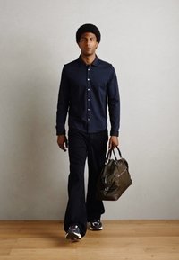 Mørkeblå skjorte med knapper, sorte bukser og sneakers, bærer en mørkeoliven grønn bag med brune detaljer. Gulv av tre.