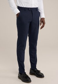 Pantalons navy sur mesure en tissu texturé, présentant un motif à carreaux subtil, une coupe ajustée et des poches latérales. Des chaussures noires complètent le look.