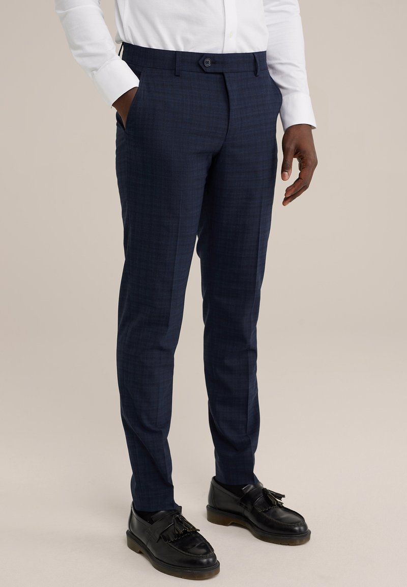 Pantalons navy sur mesure en tissu texturé, présentant un motif à carreaux subtil, une coupe ajustée et des poches latérales. Des chaussures noires complètent le look.