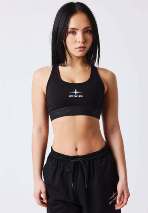 Mujer joven con cabello negro liso, vistiendo un sujetador deportivo negro y pantalones negros con cordón, sobre un fondo liso y claro.