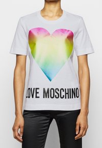 Bílý bavlněný tričko s velkým, barevným srdcovým motivem a výrazným černým textem „LOVE MOSCHINO“ na dolní části.