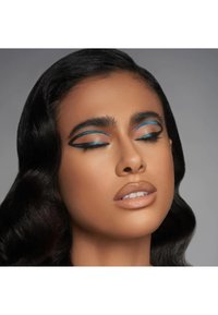 Look trucco caratterizzato da un eyeliner blu audace con dettagli neri, pelle liscia e labbra nude lucide, completato da capelli scuri e ondulati.