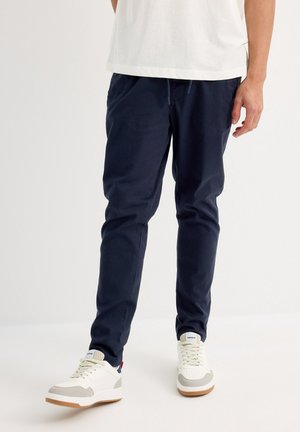 Pantalones de tiro ajustable de color navy, hechos de material ligero, de corte ajustado, con bolsillos laterales y una textura suave, combinados con zapatillas blancas.
