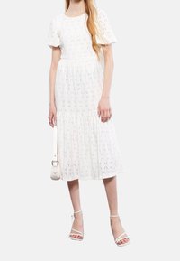 Kobieta w białej, fakturowanej sukience midi z bufiastymi rękawami, trzymająca białą torebkę i nosząca białe sandały na obcasie.