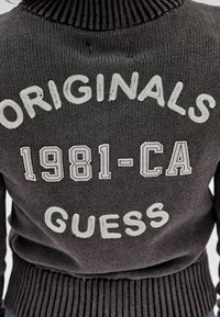 Szary sweter z prążkowanym kołnierzem i dołem, z białym napisem "ORIGINALS 1981-CA GUESS" w pogrubionej czcionce.