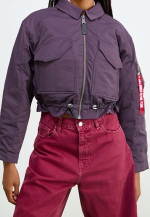 Person iført en kort lilla lynlåstrøje med forlommer og røde højtaljede bukser, stående foran en ensfarvet baggrund.