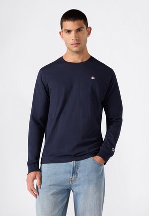 Mann trägt ein marineblaues langärmeliges Champion-Shirt und hellblaue Jeans, steht mit einer Hand in der Tasche vor einem schlichten Hintergrund.