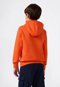 Pomarańczowy hoodie z przednią kieszenią, ściągaczami przy mankietach i kapturem ze sznurkiem. Noszony z ciemnoniebieskimi spodniami typu cargo, prezentowany z tyłu.