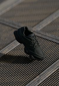 Shoe atlético preto feito de malha e materiais sintéticos, apresentando um cabedal texturizado, colarinho acolchoado e uma sola de borracha com acentos curvados.
