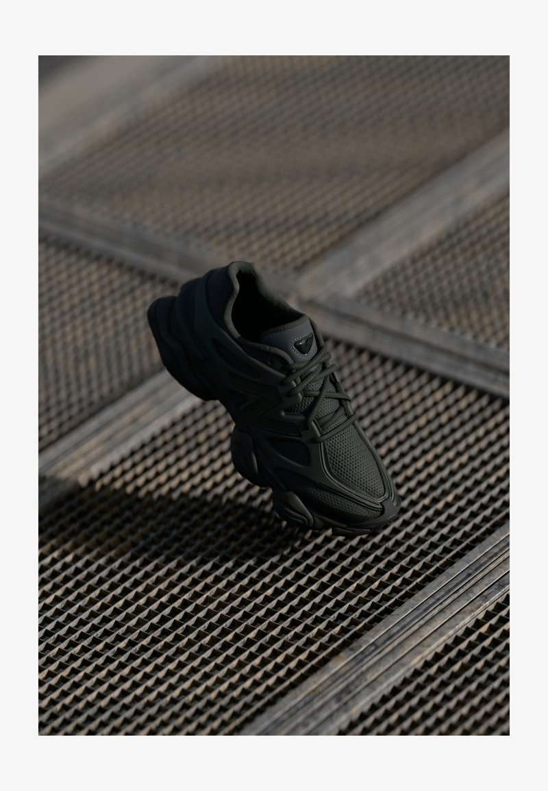 Fekete sportcipő, amely hálós és szintetikus anyagokból készült, texturált felsőrésszel, párnázott bokarészsel és ívelt díszítéssel ellátott gumitalppal.