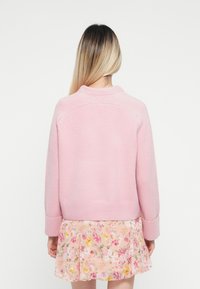Žena s dlhými blond vlasmi, oblečená v svetloružovom svetri a plisovanej sukni s kvetinovým vzorom, obrátená chrbtom na jednoduchom bielom pozadí.