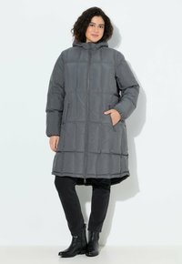 Grau gepufferte Jacke mit Kapuze, mit horizontalem Steppmuster, Reißverschluss und zwei vorderen Taschen. Kombiniert mit schwarzen Jeans und Stiefeln.